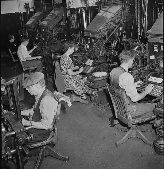 1942 linotype operators. Five people seated at large machines with complex keyboards.