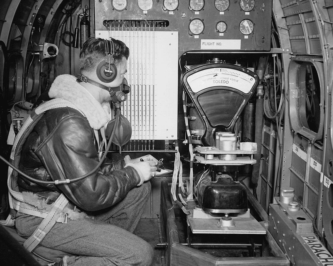 The interior of a modified B-29 bomber used to determine what conditions cause ice to form on wings and aircraft surfaces. 
An investigator wearing headphones, an oxygen mask, a fur-lined leather jacket, and stylish patterned pants is seated in front of scale. 
There are also several dial gauges.